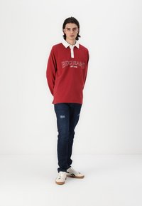 Rød langermet polo skjorte med hvit krage og teksten "BDG JEANS EST1998", kombinert med mørkeblå slitte jeans og hvite sneakers.