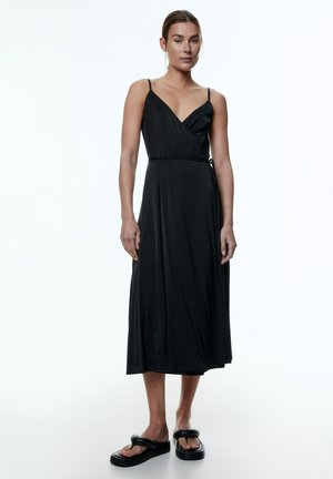 Schwarzes Wickelkleid mit tiefem V-Ausschnitt und dünnen Trägern, aus einem glatten, leichten Stoff, der bis zur Mitte der Waden reicht. Schwarze flache Sandalen.