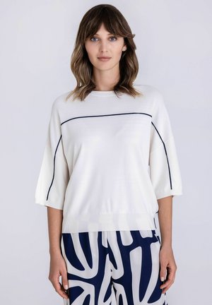 Vrouw met halflang bruin haar, gekleed in een wit bovenstuk met zwarte biezen en een broek met een abstract marineblauw-wit patroon.