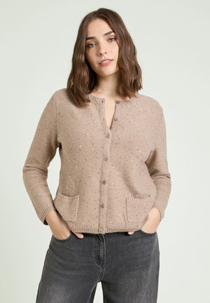 Young woman with brown hair wearing a beige buttoned cardigan with small sequins and dark gray jeans, hands in pockets.