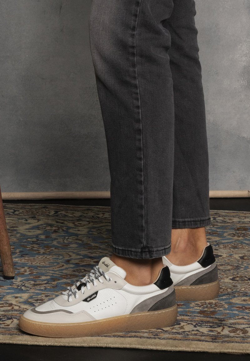 Sneaker bianche in pelle con accenti grigi e neri, caratterizzate da una finitura testurizzata, punta rotonda e lacci a contrasto. Suola in gomma con un leggero plateau.