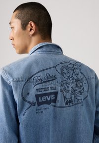 Lys blå denimjakke med et mørkt grafisk trykk av Toy Story karakterer på ryggen, med "Levi's" logo og Western-mote merkevare.
