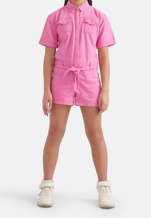 Rosa denim kort jumpsuit med korte ærmer, to brystlommer, talje med bælte og knaplukning. Kombineret med hvide høj-top sneakers.