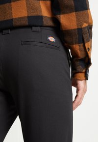 Musta housut, joissa on sileä pinta, takatasku ja näkyvä Dickies-logo. Yhdistetty oranssi-mustaraidalliseen paitaan.