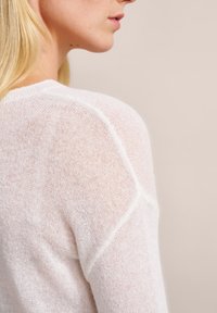 Weißer Strickpullover mit leichter, durchsichtiger Textur, der ein Raglanärmel-Design und ein dezentes Muster aufweist. Nahaufnahme im Schulterbereich.