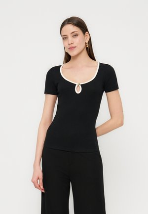 Kobieta o długich brązowych włosach nosi dopasowaną, czarną bluzkę z krótkim rękawem, z białym obszyciem i wycięciem typu keyhole, połączoną z czarnymi spodniami na jednolitym tle.