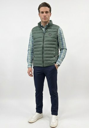 Hombre de pie vestido con un chaleco acolchado verde sobre una camisa de cuadros azul, pantalones azul marino, zapatillas blancas y un reloj de pulsera contra un fondo liso.