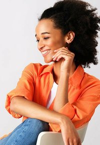 Camicia arancione con bottoni, top bianco e jeans blu. Donna con capelli ricci che appoggia il mento sulla mano, sorridendo. Sfondo semplice e neutro.