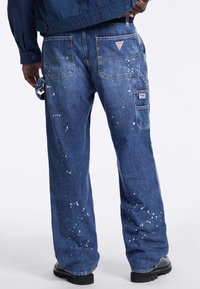 Pantaloni cargo in denim blu con una vestibilità ampia, caratterizzati da dettagli di schizzi di vernice, molteplici tasche e un'etichetta logo sulla tasca posteriore destra.