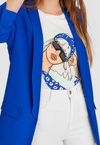 Blå blazer med en slät textur, tillsammans med en vit t-shirt som har en färgglad grafik av en kvinna med solglasögon och ett blått hårband.