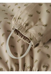 Tela beige con un patrón repetido de conejitos; cuenta con frunces elásticos y un bucle de plástico. Textura suave con un brillo sutil.