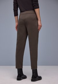 Bruine taps toelopende broek met een gladde textuur, voorzien van twee achterzakken, gecombineerd met zwarte enkellaarsjes, tegen een neutrale achtergrond.