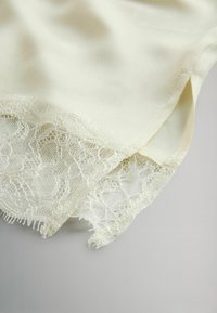 Cream-colored fabric with lace trim at the hem. The lace features floral patterns and a delicate texture, with frayed edges for detail.