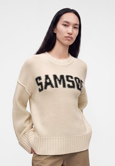 Млада жена с дълга тъмна коса носи кремава плетена блуза с черен текст "SAMSO" и кафяви панталони, стои пред едноцветен фон.
