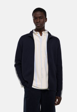 Jeune homme portant une veste zippée bleu marine avec des bandes blanches sur les manches, par-dessus une chemise à rayures crème et bleu clair, debout devant un fond uni.