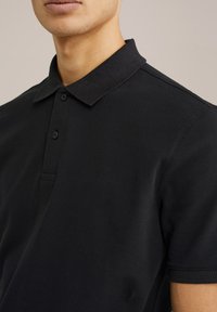 Musta poolpaita, joka on valmistettu teksturoidusta kankaasta, jossa on klassinen kaulus, kaksinappinen lista ja lyhyet hihat.