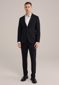 Tumesinine ülikond, millel on lõigatud kraed, kaks nuppu ja küljetaskud, koos valge särgiga ja mustade kingadega, seismas neutraalse tausta ees.