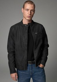 Chaqueta de cuero negra con cuello alzado, cremallera frontal y acabado texturizado. Presenta un bolsillo en el pecho con cremallera y se lleva sobre una camiseta negra.