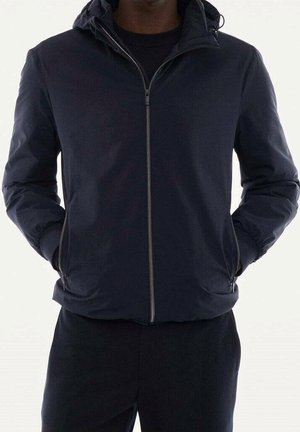 Uomo che indossa una giacca nera con zip e cappuccio, con le mani nelle tasche laterali, mostrato dal collo ai fianchi su uno sfondo semplice.