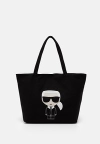 Černá taška přes rameno s dvěma uchy, která zobrazuje minimalistický karikaturu osoby s bílými vlasy, tmavými slunečními brýlemi a černým oblekem s kravatou.