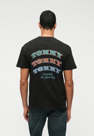 T-shirt de algodão preto com texto "TOMMY" multicolorido em várias fontes nas costas, acompanhado de "JEANS New York City" em azul claro.