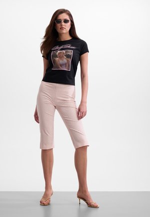 Femme portant un t-shirt noir graphique avec une photo rose, pantalon capri rose à carreaux, lunettes de soleil et sandales à talons beiges, debout.