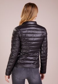Sort, puffy jakke med vandret syninger, blank finish og høj krave, parret med grå tætsiddende jeans.