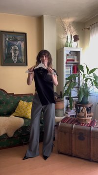 Zwarte mouwloze top aan een witte hanger, met een getailleerd ontwerp. Model draagt grijze wijde broek en zwarte puntige schoenen.