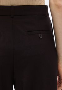 Pantalon marron sur mesure avec une texture lisse, doté d'une ceinture plate et de deux poches arrière avec fermetures à boutons.