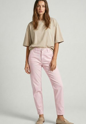 Woman standing, wearing a loose beige t-shirt, light pink pants, and beige loafers against a plain light background.