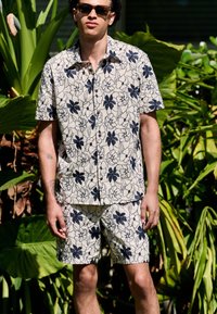 Kurzärmeliges Hemd mit Blumenmuster und passende Shorts in hellbeige mit dunkelblauen Blumen. Das Material wirkt leicht; das Design ist lässig.