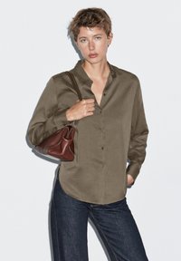 Chemise olive en tissu doux, à manches longues retroussées et à la coupe décontractée, associée à un sac en cuir marron. Un jean foncé vient compléter le look.