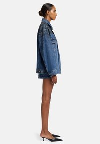 Giubbotto di jeans con una vestibilità rilassata, colore blu scuro, abbellito da borchie nere sulle spalle e due tasche frontali; indossato con una gonna coordinata.