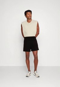 Bézs ujjatlan felső, fekete rövidnadrág húzózsinórral és világosszürke sportcipő. Egyszerű kialakítás laza szabással és minimális díszítéssel.