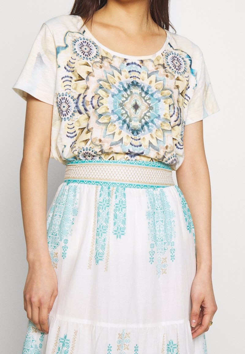 Femme portant un haut à manches courtes avec un motif de mandala bleu et beige, rentré dans une jupe blanche ornée de motifs géométriques teal et beige et d'une ceinture à motifs.