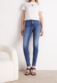 Bílý zkrácený tričko s černým logem, modré skinny jeans se zeslabeným praním a černé sandály na vysokém podpatku s pásky. Minimalistický design.