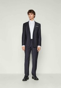 Jovem em um fato escuro bem ajustado, camisa branca, sapatos pretos, contra um fundo claro e simples.