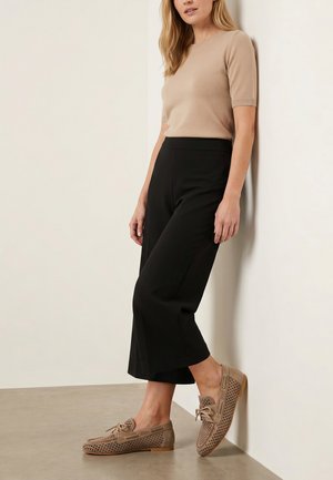 Kvinde, der læner sig op ad en væg, iført en beige kortærmet top, sorte vidde cropped bukser og taupe perforerede loafers med snørebånd.