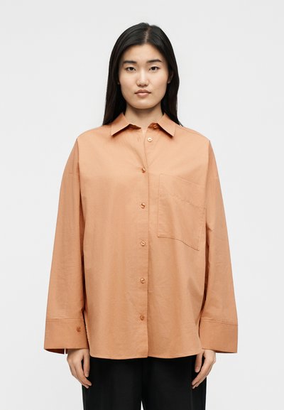 Žena s dlhými tmavými vlasmi, ktorá má na sebe voľnú, veľkú svetlohnedú košeľu s dlhými rukávmi a predným náprsným vreckom.