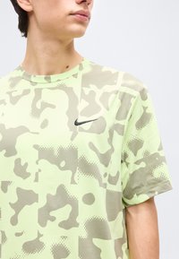 Νεαρό άτομο που φοράει ανοιχτό πράσινο και γκρι T-shirt με σχέδιο καμουφλάζ της Nike, στραμμένο παράλληλα σε απλό φόντο.