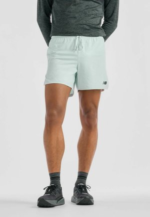 Person trägt hellblaue Sportshorts, schwarze und graue Laufschuhe und ein dunkles Langarmshirt vor einfachem Hintergrund.
