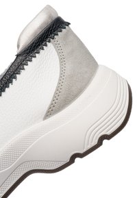 Primer plano del talón de una zapatilla blanca con cuero texturizado, detalles en gamuza gris, ribete de costura negra y suela gruesa de goma blanca con dibujo marrón.