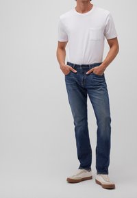 Hombre con camiseta blanca con bolsillos, jeans azules y zapatillas beige de pie con las manos en los bolsillos frente a un fondo sencillo.