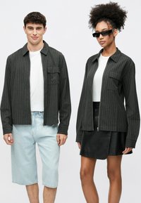 Dwie osoby noszą czarne koszule w prążki: jedna z suwakiem i naszywanymi kieszeniami, w połączeniu z jasnoniebieskimi szortami, a druga z czarną skórzaną spódnicą.