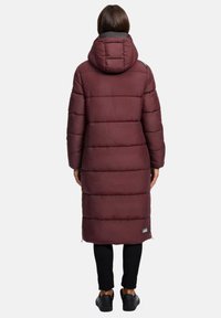 Person som står med ryggen mot kameran iförd en lång burgunderröd vadderad kappa med huva, svarta byxor och svarta sneakers mot en enkel bakgrund.