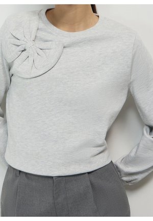 Vrouw draagt een lichtgrijze top met lange mouwen en een grote stoffen strik op de linkerschouder, ingestopt in een hooggetailleerde donkergrijze broek.