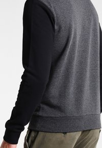 Grå och svart sweatshirt med texturerad kropp, svarta ärmar, ribbade muddar och fåll. Har rund halsringning och sidofickor.