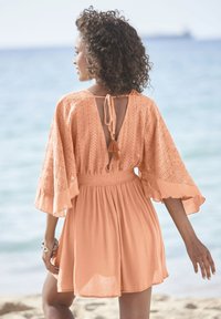 Perzikkleurige jurk met een gestructureerde lijfje, open rug, uitgezette mouwen en een geraffineerde taille. Bevat een striksluiting en een vloeiende rok.