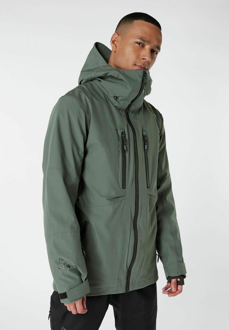 Giacca impermeabile verde con colletto alto, zip anteriore e molteplici tasche con zip. Presenta una texture liscia e polsini regolabili.