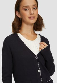 Granatowy kardigan z miękkiej dzianiny z falistym kołnierzem i białym wykończeniem, z dwiema przednimi kieszeniami i zapięciem na guziki.